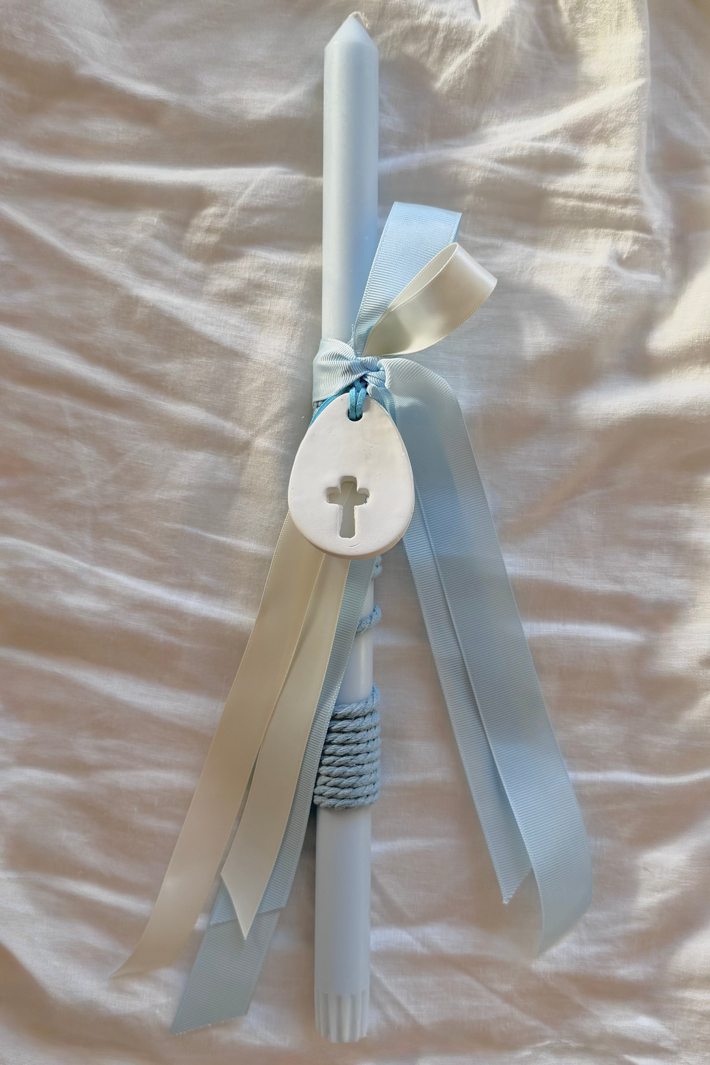 White candle with blue ribbons and a cross charm on a beige fabric background