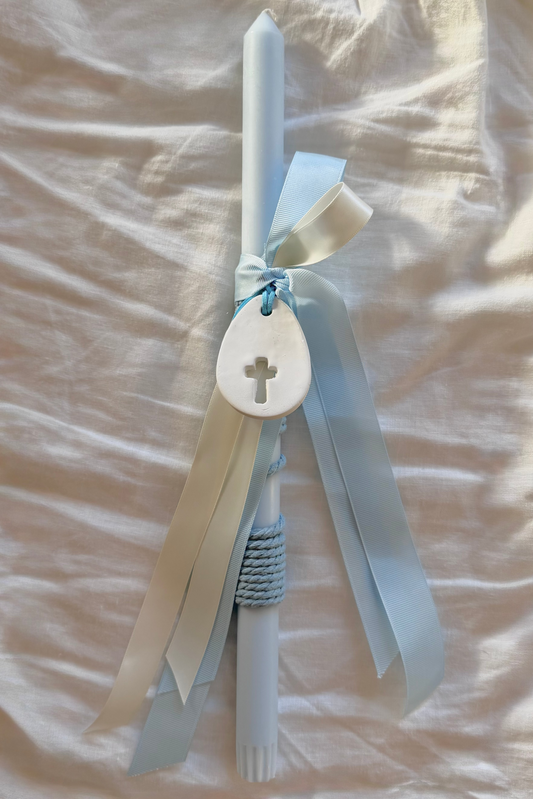 White candle with blue ribbons and a cross charm on a beige fabric background